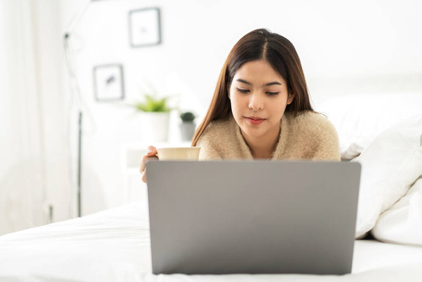 Młoda uśmiechnięta szczęśliwa piękna azjatycka kobieta relaksująca się przy użyciu laptopa w sypialni w domu.Młoda kreatywna dziewczyna pracująca i pisząca na keyboard.work z koncepcji domu - Zdjęcie, obraz