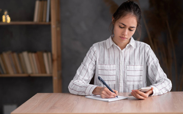 young hispanic woman freelance with smartphone chat with colleagues writing on notebook at home office. Girl student learn online, home job concept. - Foto, Bild