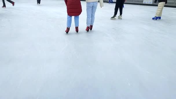 Emberek korcsolyáznak nyilvános szabadtéri jégpályán a városban télen. Az emberek korcsolyáznak a jégpályán. Lábak, hátra, jégpálya. Téli ünnepek, tevékenységek, kikapcsolódás, pihenés, sport - Felvétel, videó