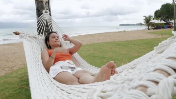 Ασιάτισσα γυναίκα χαλαρωτική και σε αιώρα κοντά στην παραλία κάτω από την καρύδα δέντρο σε διακοπές - Πλάνα, βίντεο