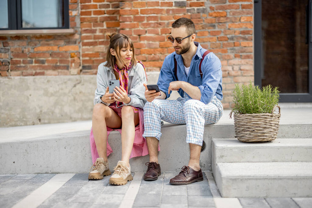 Νεαρός κομψός άντρας και γυναίκα κάθονται μαζί με έξυπνα τηλέφωνα και έχουν κάποια συζήτηση κοντά στο γραφείο σε εξωτερικούς χώρους. Σύγχρονοι hipsters κατά τη διάρκεια διάλειμμα καφέ - Φωτογραφία, εικόνα