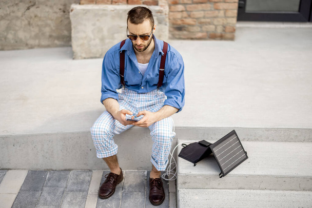 Stylish man use smart phone and charge it from portable solar panel while sitting on street outdoors. Modern sustainable lifestyle and renewable energy - Foto, Bild