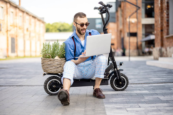 Stylish man works on laptop computer while sitting on electric scooter on a street at office district. Concept of sustainable and eco friendly lifestyle - Photo, Image