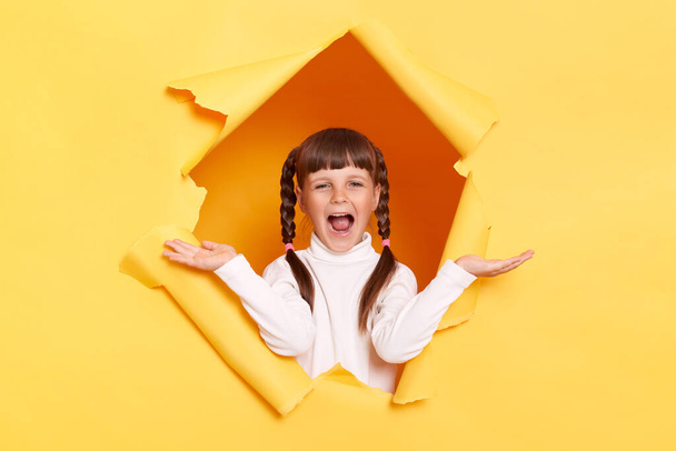 Portrait of amazed little girl with braids wearing white turtleneck posing in torn hole of yellow paper wall, standing yelling happily, celebrating her success, spreading hands. - Zdjęcie, obraz