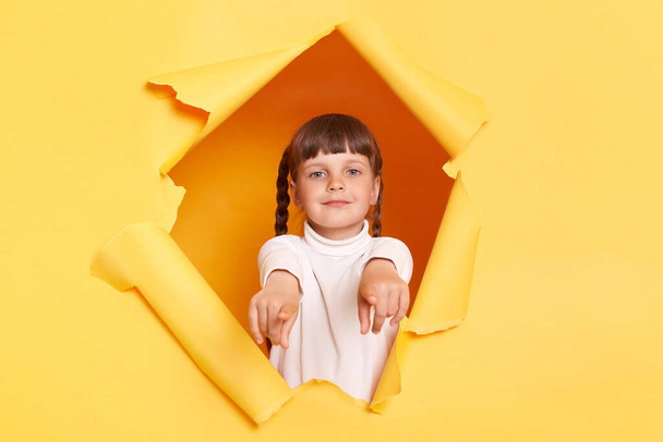 Portrait of delighted positive Caucasian little girl with braids wearing white turtleneck, pointing both index fingers to camera, choosing you, posing in torn hole of yellow paper wall. - Foto, immagini