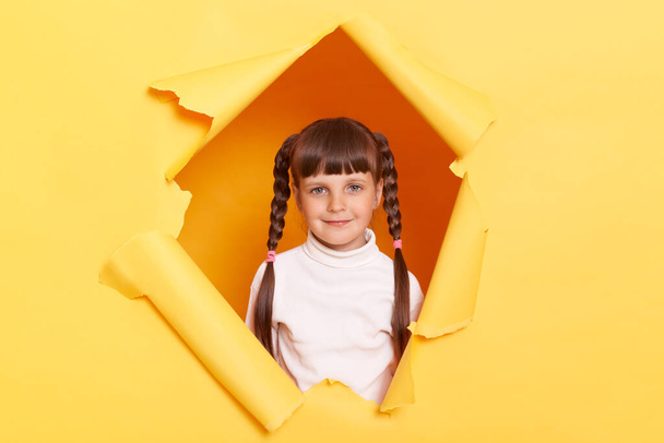 Image of satisfied adorable charming little girl with braids wearing white turtleneck posing in torn hole of yellow paper wall, looking at camera, expressing calm emotions. - Φωτογραφία, εικόνα