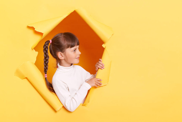 Side view of caucasian positive little girl with braids wearing white turtleneck posing in torn hole of yellow paper wall, standing looking away at advertisement area. - Valokuva, kuva