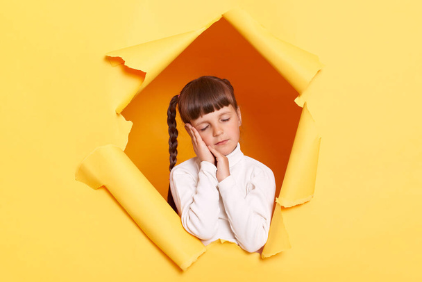 Horizontal shot of sleepy tired little girl with braids wearing white turtleneck posing in torn hole of yellow paper wall, napping while leaning on her palms, keeps eyes closed. - Zdjęcie, obraz