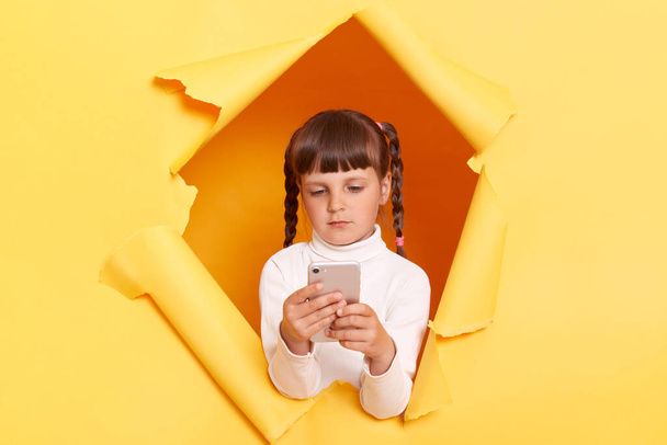 Portrait of serious concentrated little girl with braids wearing white turtleneck, holding cell phone, looking at display, using mobile phone, posing in torn hole of yellow paper wall. - Fotografie, Obrázek