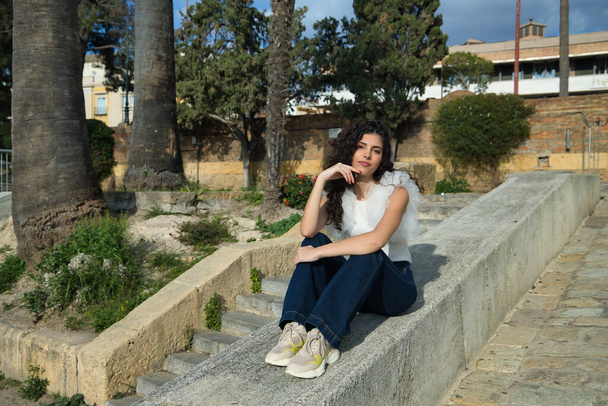 Young and beautiful woman, brunette, with curly hair and affectionate gesture, wearing a white shirt and jeans, sitting on a concrete wall. Concept beauty, fashion, model, affection, trend. - Foto, afbeelding