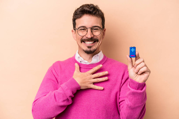 Young caucasian man holding batteries isolated on beige background laughs out loudly keeping hand on chest. - Foto, imagen