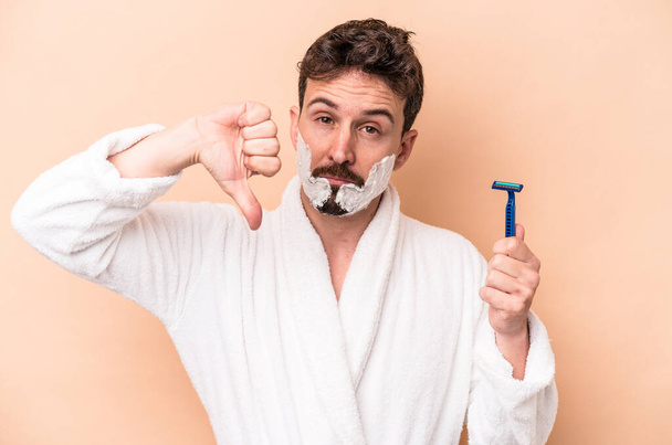 Young caucasian man wearing shaving foam and holding razor blade isolated on beige background showing a dislike gesture, thumbs down. Disagreement concept. - Foto, imagen