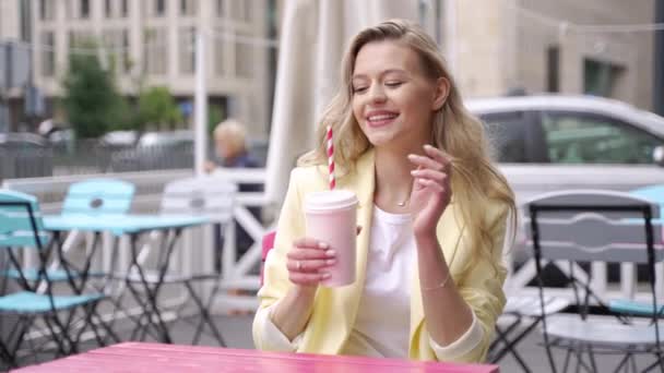 Onnellinen nuori nainen rento vaatteita hymyilee ja katselee kameraa istuessaan pöydässä kuppi vihanneksia ja hedelmiä ravistaa ulkona kahvilassa - Materiaali, video