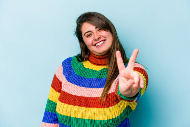 Young caucasian overweight woman isolated on blue background joyful and carefree showing a peace symbol with fingers. - 写真・画像