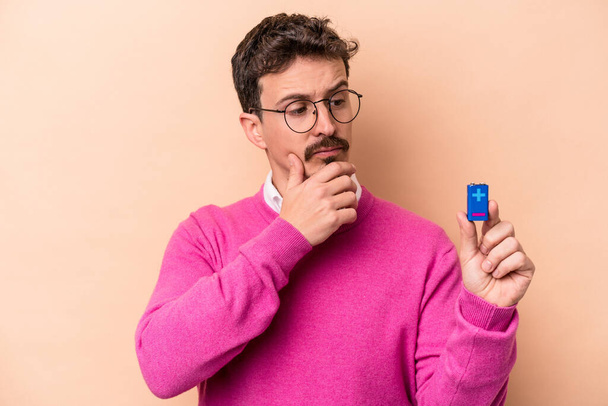 Young caucasian man holding batteries isolated on beige background looking sideways with doubtful and skeptical expression. - Φωτογραφία, εικόνα