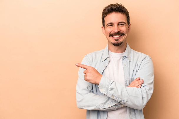 Young caucasian man isolated on beige background smiling cheerfully pointing with forefinger away. - Foto, immagini