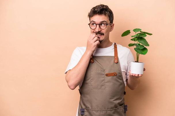 Jovem jardineiro caucasiano homem segurando uma planta isolada em fundo bege relaxado pensando em algo olhando para um espaço de cópia. - Foto, Imagem