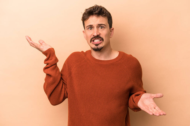 Young caucasian man isolated on beige background confused and doubtful shrugging shoulders to hold a copy space. - Foto, Imagen
