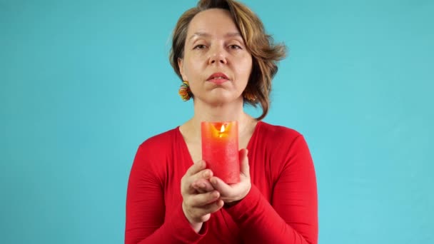 Czterdziestoletnia kobieta w czerwonym swetrze trzyma płonącą świecę na niebieskim tle. Portret dorosłej kobiety. zwolniony ruch - Materiał filmowy, wideo