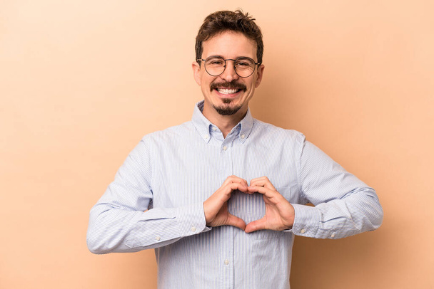 Молодий білий чоловік ізольований на бежевому фоні посміхається і показує форму серця руками
. - Фото, зображення