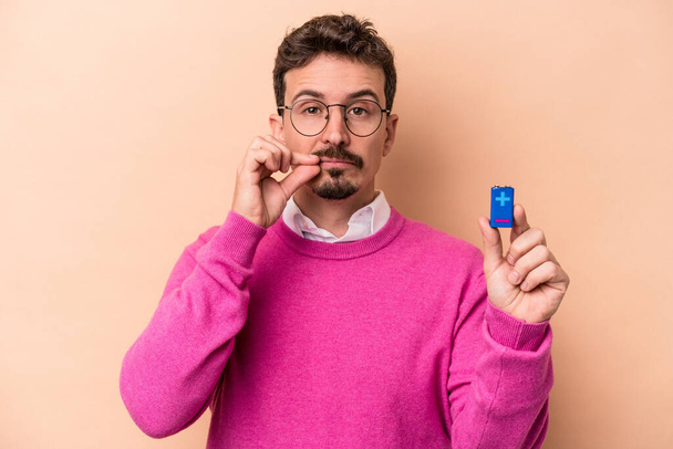 Young caucasian man holding batteries isolated on beige background with fingers on lips keeping a secret. - Fotoğraf, Görsel