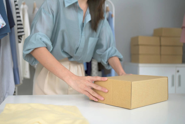 Happy Asian young woman showing a cardboard parcel box to camera, online selling or ecommerce concept. Female online seller sells a clothes via online marketplace platform. - Фото, изображение