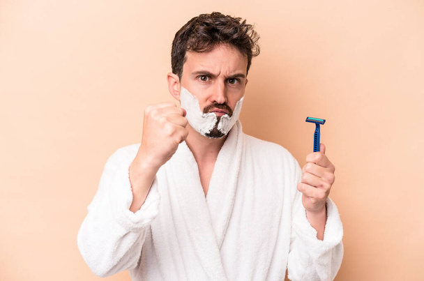 Young caucasian man wearing shaving foam and holding razor blade isolated on beige background showing fist to camera, aggressive facial expression. - Фото, зображення