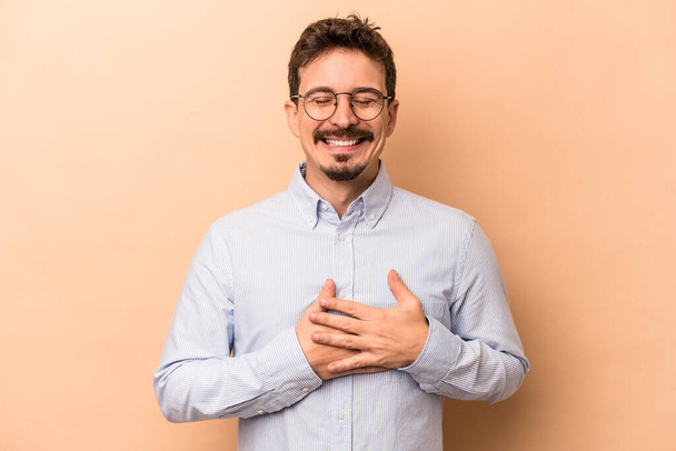 Young caucasian man isolated on beige background laughing keeping hands on heart, concept of happiness. - Foto, Imagem