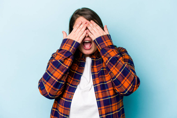 Jonge blanke vrouw met overgewicht geïsoleerd op blauwe achtergrond bedekt ogen met handen, glimlacht breed te wachten op een verrassing. - Foto, afbeelding