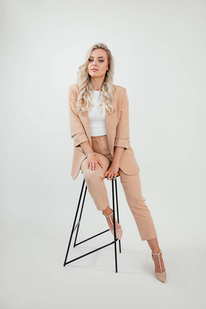 Blonde long haired cute business Woman wearing stylish formal wear beige suit on white background, sitting on chair, studio shot, look book  - Photo, Image