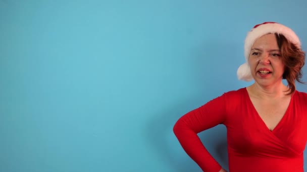 Una mujer adulta con una chaqueta roja y un sombrero de Año Nuevo sobre fondo azul. La mujer suspira. Retrato en cámara lenta - Metraje, vídeo