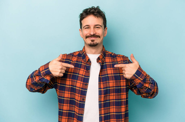 Young caucasian man isolated on blue background person pointing by hand to a shirt copy space, proud and confident - Foto, imagen