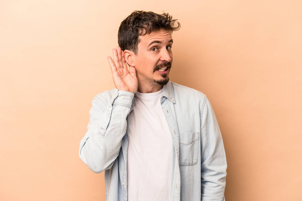 Young caucasian man isolated on beige background trying to listening a gossip. - Photo, Image