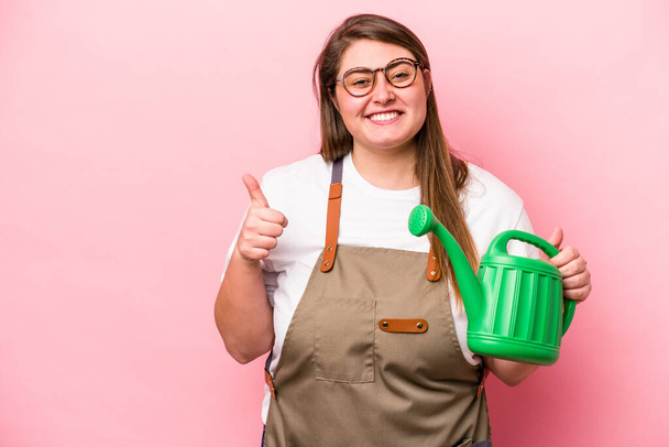 Young gardener caucasian overweight woman holding watering can isolated background smiling and raising thumb up - Foto, Imagen