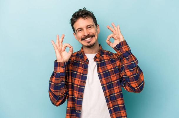 Young caucasian man isolated on blue background cheerful and confident showing ok gesture. - Photo, Image