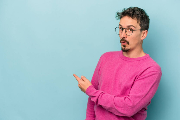Young caucasian man isolated on blue background smiling and pointing aside, showing something at blank space. - Foto, Imagen
