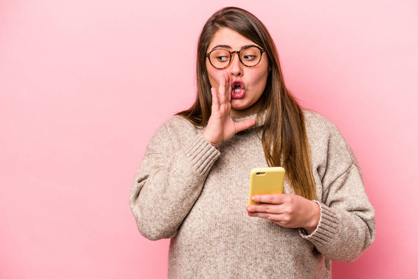 Fiatal fehér túlsúlyos nő kezében egy mobiltelefon elszigetelt rózsaszín háttér azt mondja, egy titkos forró fékezés hír, és félrenéz - Fotó, kép