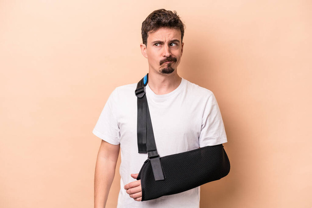 Young caucasian man with broken arm isolated on beige background confused, feels doubtful and unsure. - Photo, Image