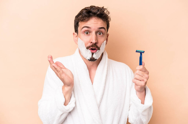 Jeune homme caucasien portant de la mousse à raser et tenant lame de rasoir isolé sur fond beige surpris et choqué. - Photo, image
