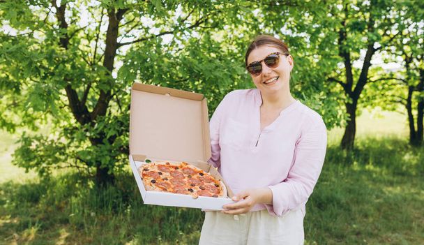 Счастливая молодая женщина держит горячую пиццу в коробке на фоне природы. Привлекательная девушка чувствует себя голодной и любит вкус. Вся пицца для меня. - Фото, изображение