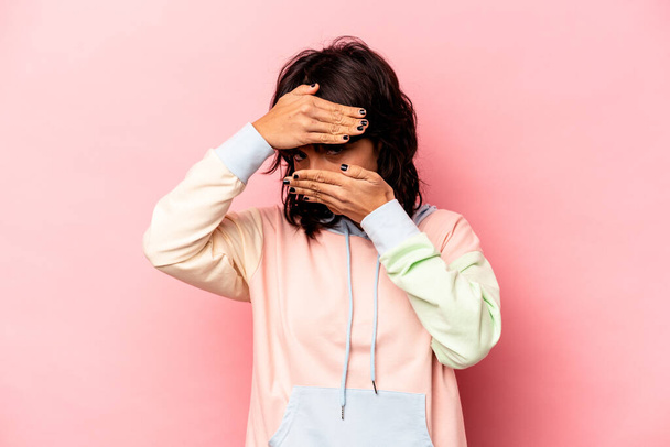Młoda Latynoska kobieta odizolowana na różowym tle mruga do kamery przez palce, zakłopotana przykrywająca twarz. - Zdjęcie, obraz