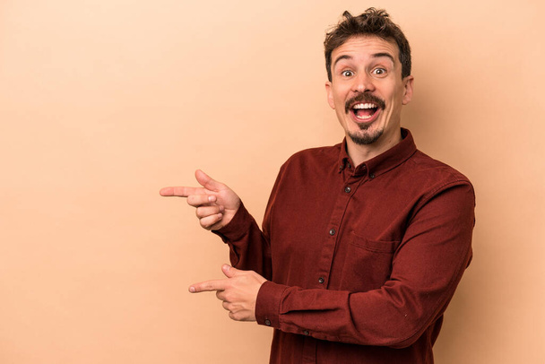 Young caucasian man isolated on beige background excited pointing with forefingers away. - Fotó, kép