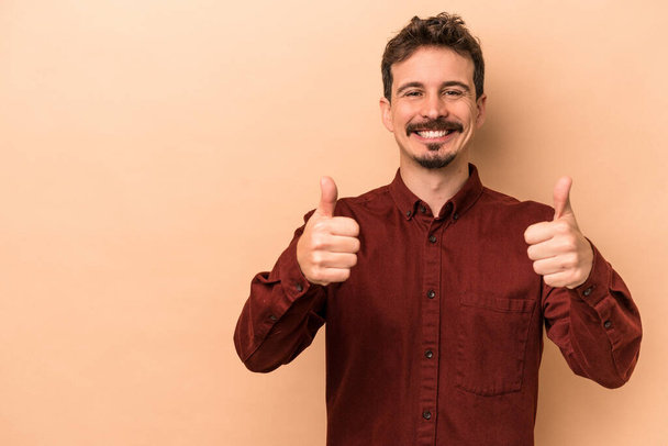 Young caucasian man isolated on beige background smiling and raising thumb up - Photo, Image