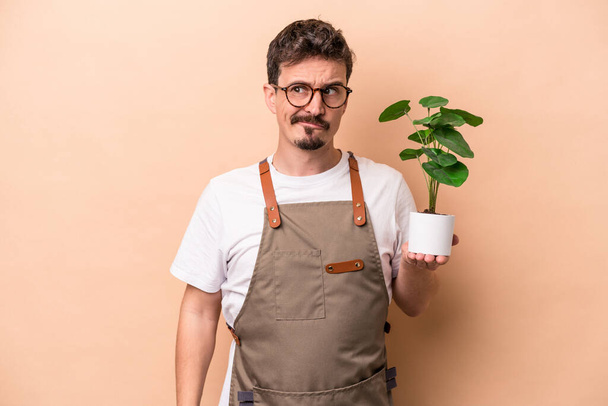 Joven jardinero caucásico hombre sosteniendo una planta aislada sobre fondo beige confundido, se siente dudoso e inseguro. - Foto, imagen