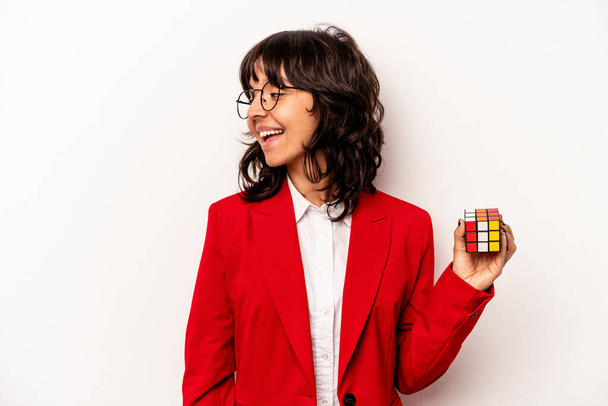 Νεαρή επιχειρηματίας κρατώντας κύβο Rubiks απομονωμένη σε λευκό φόντο κοιτάζει πέρα χαμογελαστός, χαρούμενος και ευχάριστος. - Φωτογραφία, εικόνα