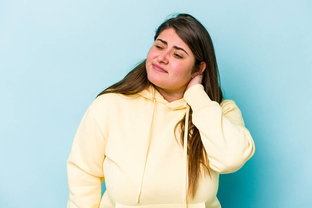 Молодая белая женщина с избыточным весом, изолированная на синем фоне, испытывающая боль в шее из-за стресса, массирующая и трогающая ее рукой. - Фото, изображение