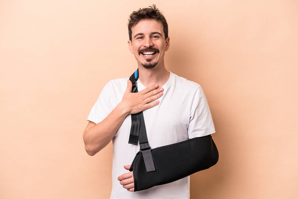 Young caucasian man with broken arm isolated on beige background laughs out loudly keeping hand on chest. - Foto, Imagen