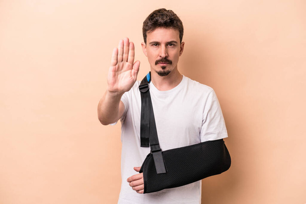 Young caucasian man with broken arm isolated on beige background standing with outstretched hand showing stop sign, preventing you. - Photo, Image