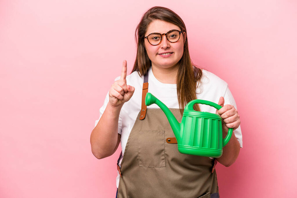 Young gardener caucasian overweight woman holding watering can isolated background showing number one with finger. - Photo, Image