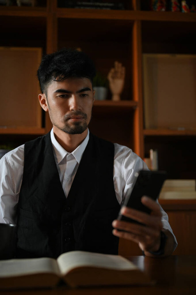 Porträt eines professionellen asiatischen Geschäftsmannes oder männlichen Unternehmers an seinem Bürotisch, der ein Smartphone benutzt, um mit jemandem zu chatten. - Foto, Bild